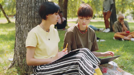 Zwei-Mädchen-Sitzen-Mit-Laptop-Im-Park-Und-Unterhalten-Sich