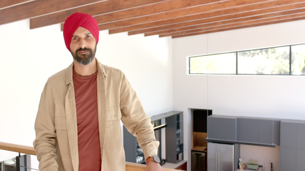 Uomo in piedi in una casa moderna, che indossa un turbante rosso e abiti casual.