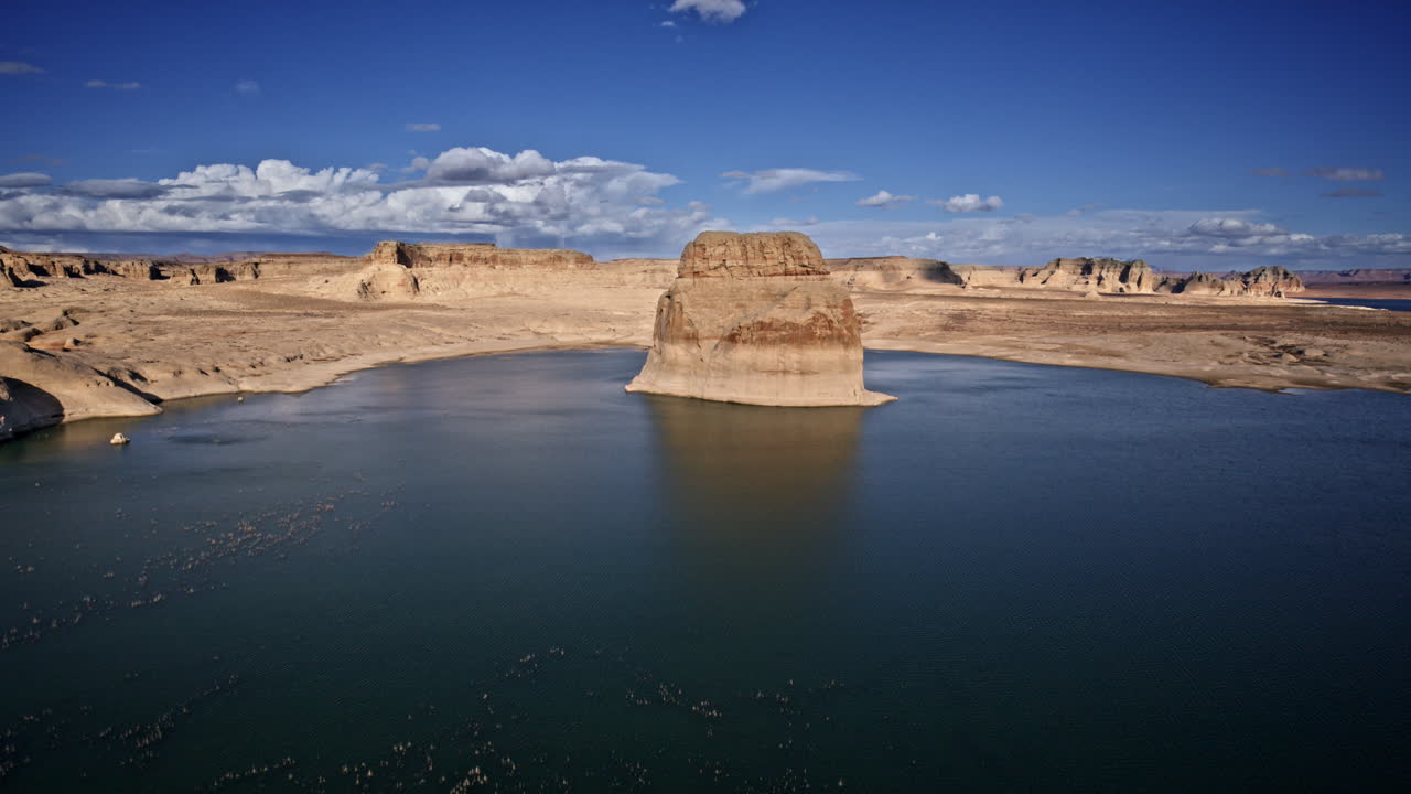 Drone shot showcasing a massive rock formation rising from Lake Powell, framed by the rugged desert canyons near Page, Arizona.