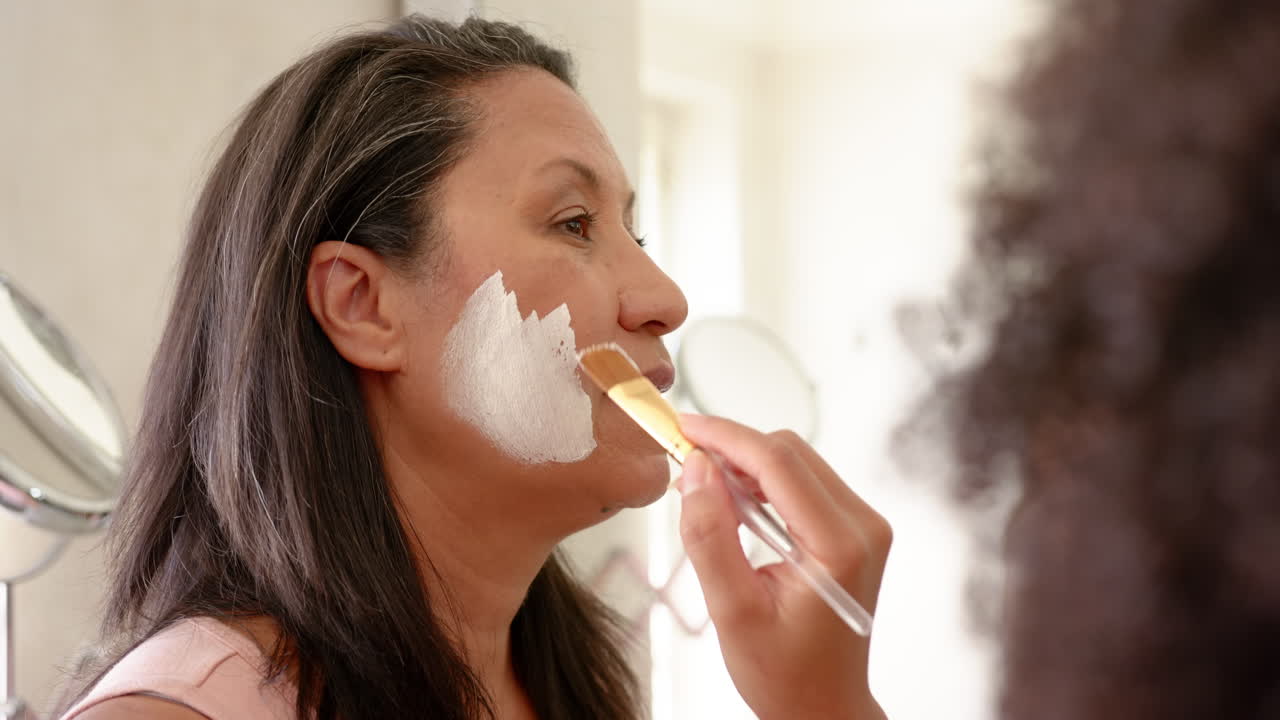 Applying face mask, woman receiving skincare treatment with brush at home