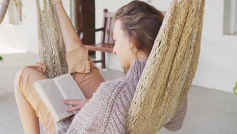 mujer caucásica sonriente acostada en una hamaca leyendo un libro en la terraza