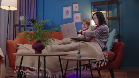 madre y niño pequeño hija niño estudio en la computadora portátil, viendo películas divertidas, relajándose, acostado en el sofá
