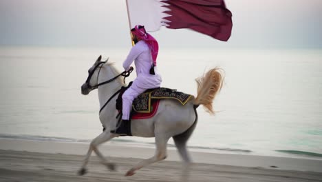 um cavaleiro andando a cavalo correndo e segurando a bandeira do qatar perto do mar-3