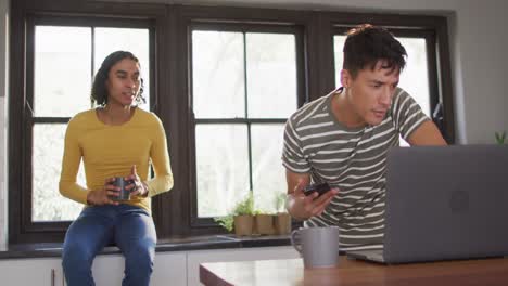 Happy-diverse-male-couple-drinking-coffee-and-using-laptop-in-kitchen