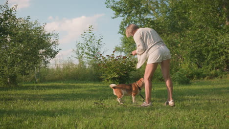 dog owner dancing joyfully with her dog, who jumps excitedly towards her as she offers treat, happening in lush green garden under sunny weather surrounded by trees