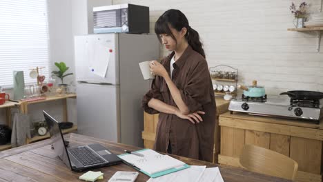 asian woman creative worker sipping tea and looking at computer in contemplation with an arm on chest is bending over to type on keyboard while working from home