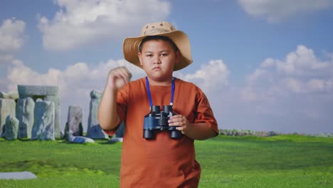 asian boy with a hat showing thumbs down gesture after looking through the binoculars. boy researcher examines something while traveling in stonehenge, travel tourism adventure concept