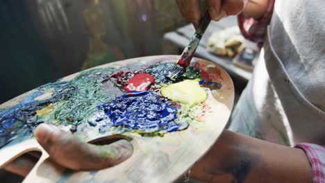 Hands-of-biracial-female-artist-mixing-paints-on-palette-using-brush-in-sunny-garden,-slow-motion