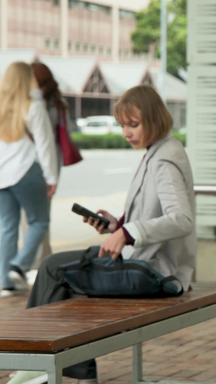 Vertical video: Glancing up, woman in grey blazer taking from blue bag, leaving bench with phone