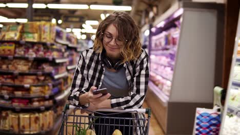 Concepto-De-Compras,-Tecnología,-Venta,-Consumismo-Y-Personas:-Mujer-Con-Teléfono-Inteligente-Y-Auriculares-En-El-Cuello-Caminando-En-El-Supermercado