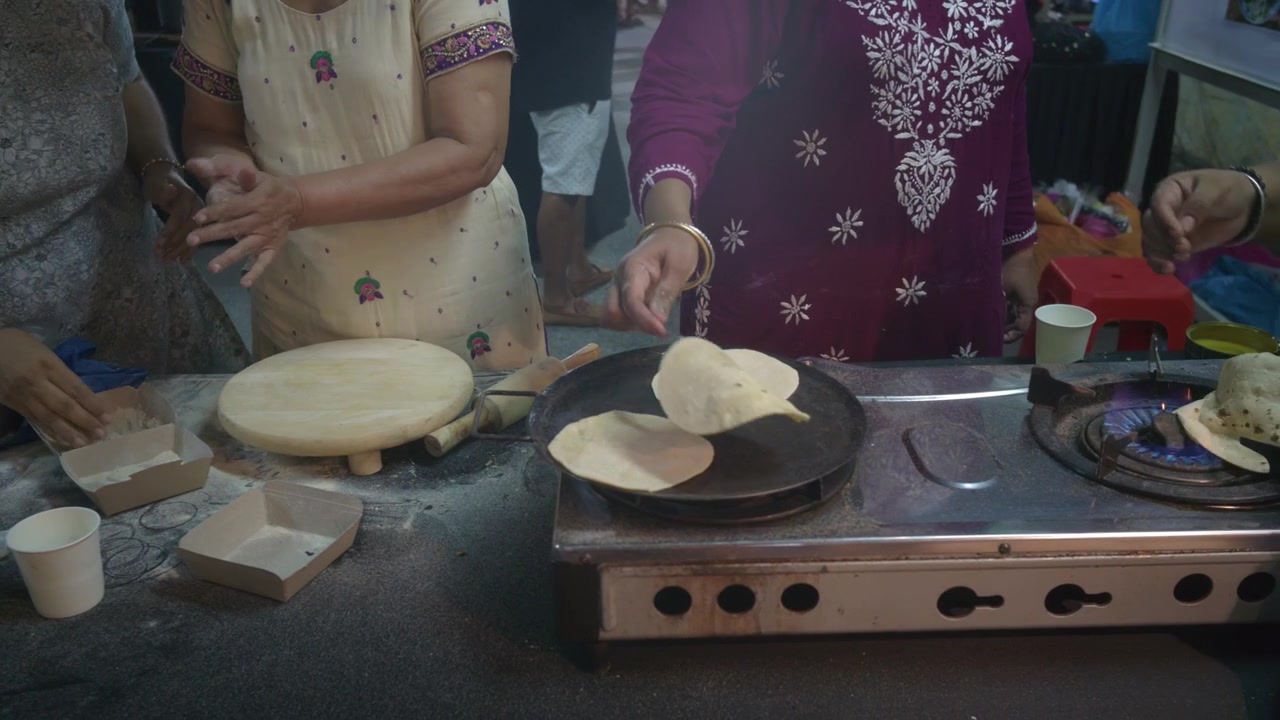 Rekaman makanan gaya dokumenter yang menunjukkan wanita menyiapkan roti pipih segar atau chapati di atas wajan panas di atas kompor gas. Adegan ini meliputi tepung, papan penggiling, penggilas adonan, peralatan masak