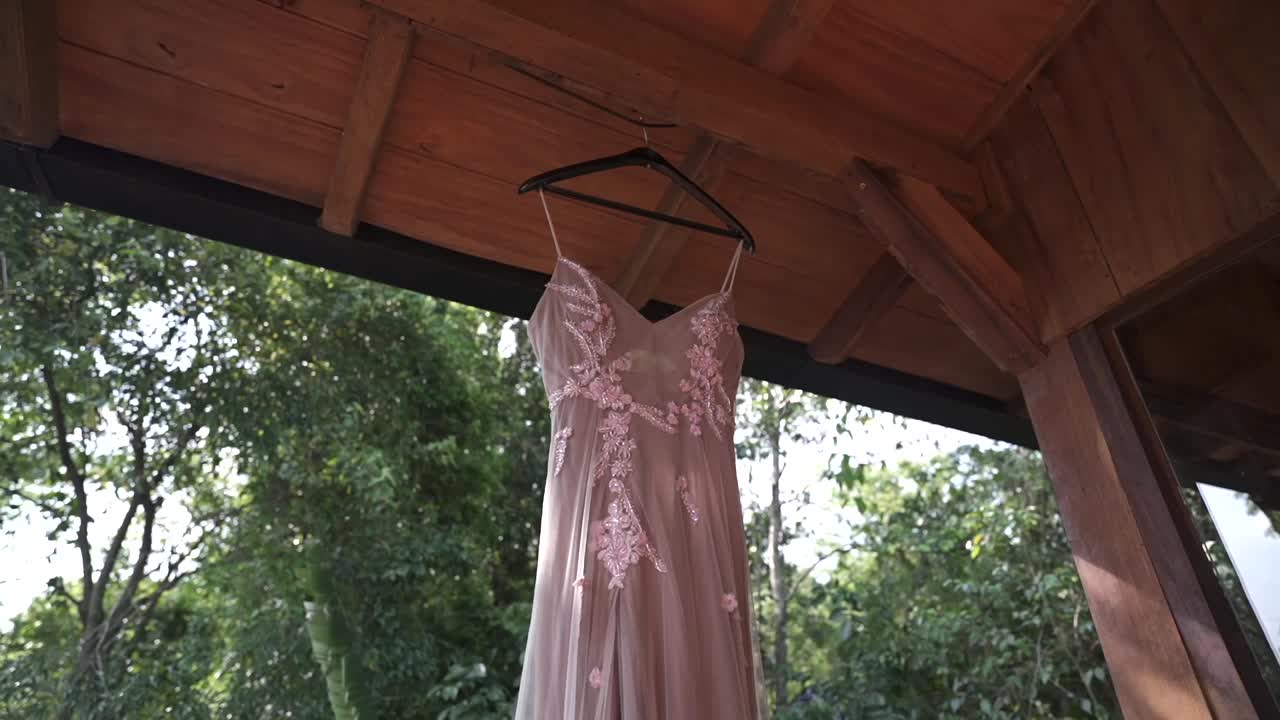 Orbiting shot in low angle around a wedding dress hanging under a wooden structure, with soft natural light and green vegetation in the background