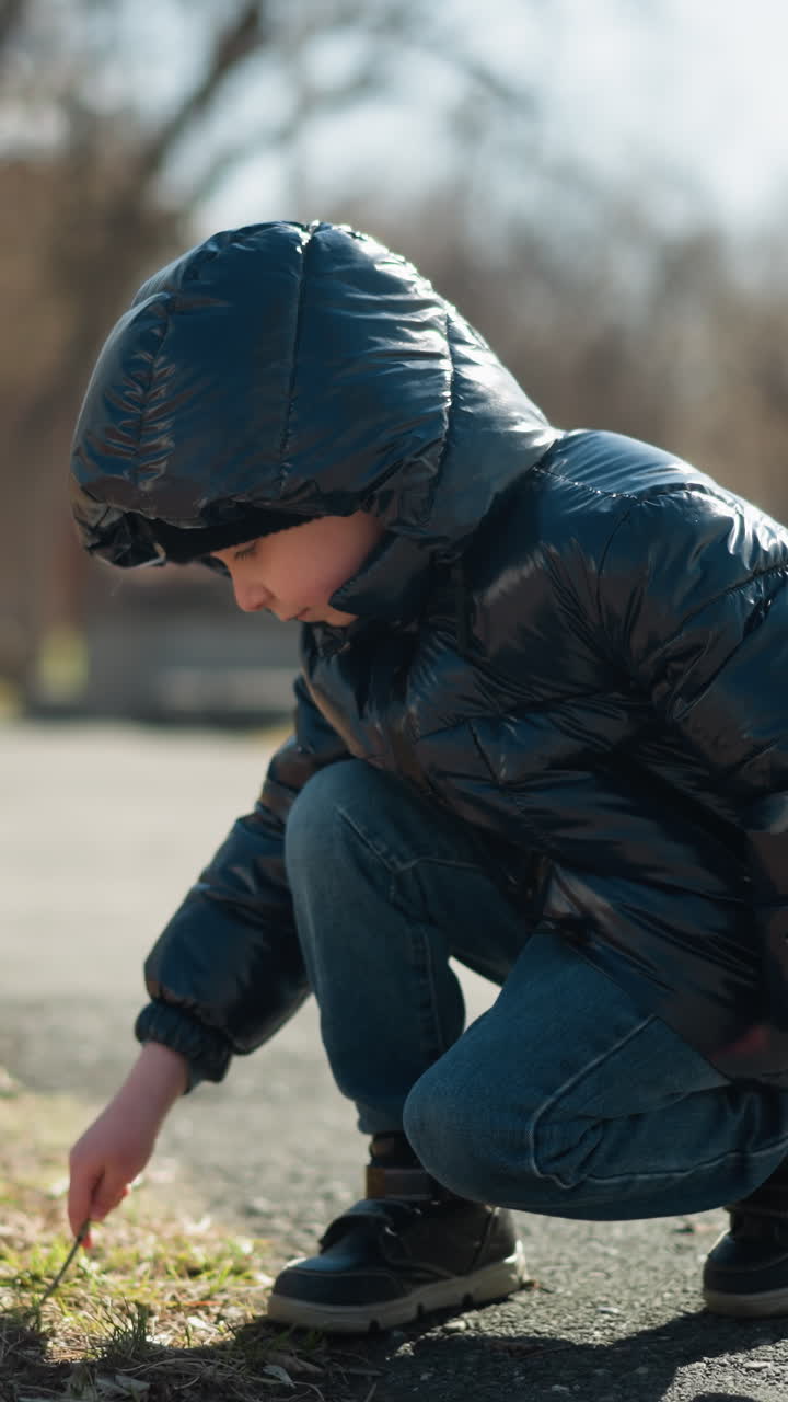 반이는 검은색 재, 파란색 청바지, 부츠를 입은 어린 소년은 잎이 없는 나무의 전망과 함께 바닥에 막대기를 가지고 놀고 있는 포장된 길에 누워 있습니다.