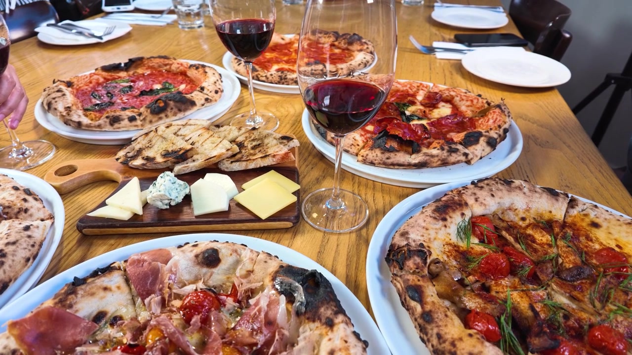 Draufsicht auf verschiedene Pizzen, eine Käseplatte und Rotwein auf einem Holztisch.