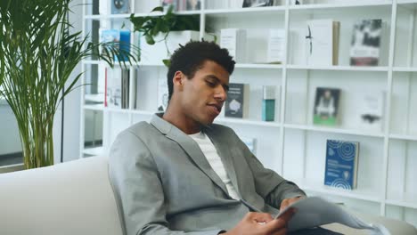 black businessman reading paper on sofa. adult african-american man in suit sitting on sofa in office and looking through paper documents