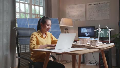 smile asian woman typing on a laptop next to the model of a small house with solar panel and wind turbine at the office