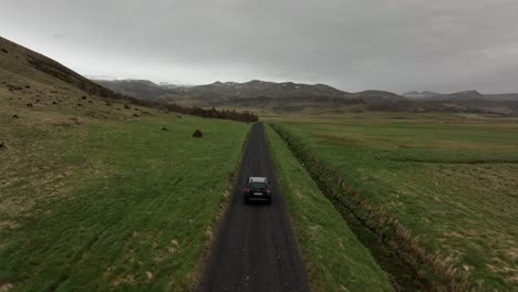 vista aérea de un automóvil que viaja por un camino estrecho a través de campos verdes en el campo islandés, rodeado de montañas lejanas bajo un cielo nublado