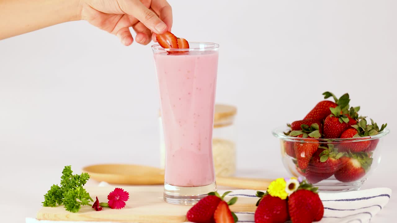 Una mano appoggia una fragola su un frullato rosa in un bicchiere, illuminazione brillante da studio, ambiente da cucina pulito