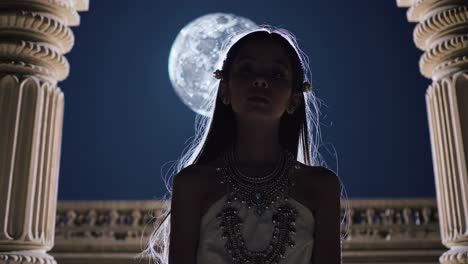 female student practicing tranquil meditation, wearing ornate cultural attire, seated between weathered stone columns under luminous moonlight, eyes gently closed