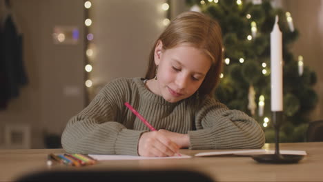 girl in green sweater writing a letter of wishes sitting at a table in a room decorated with a christmas tree