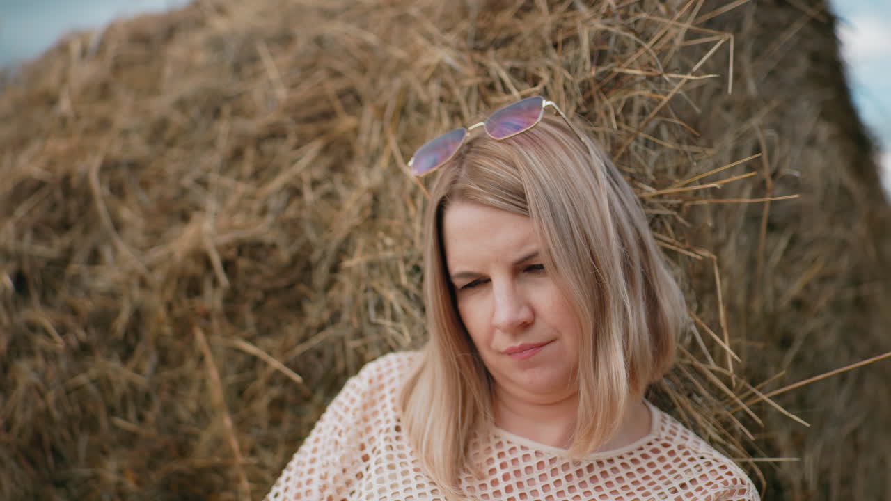 женщина, опирающаяся на шлейф сена, в солнцезащитных очках на голове и сетчатке, в спокойной сельской местности с сухой травой, голубым небом и расслабленным выражением лица