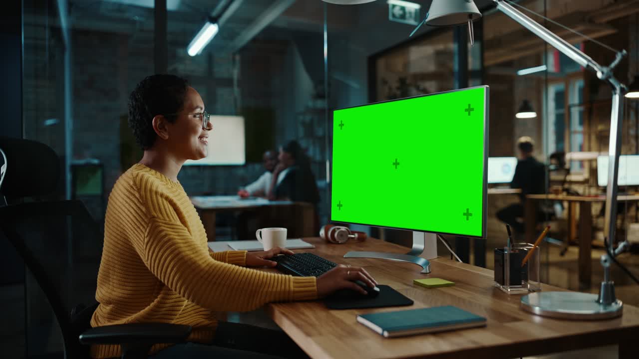 joven especialista multiétnico trabajando en una computadora de escritorio con pantalla verde simulacro de visualización en la oficina creativa. hermosa gerente femenina diversa con cabello corto y gafas está usando un jumper amarillo.