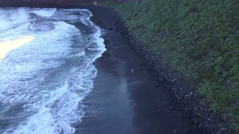 Isolated-man-from-afar-walking-forwards-at-volcanic-black-sand-coastline-in-La-Palma-island-while-waves-break