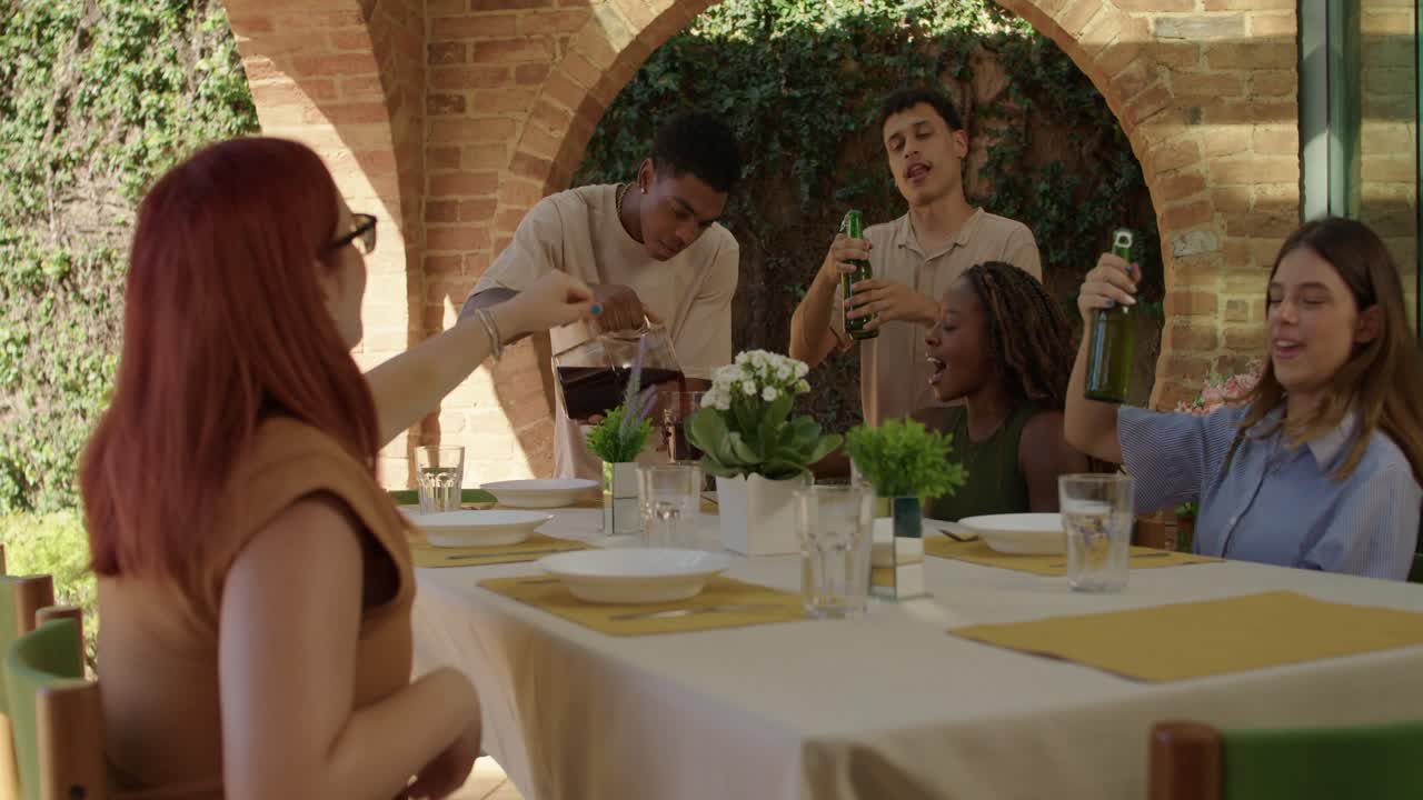A diverse group of young friends enjoys an outdoor meal and drinks on a sunny day