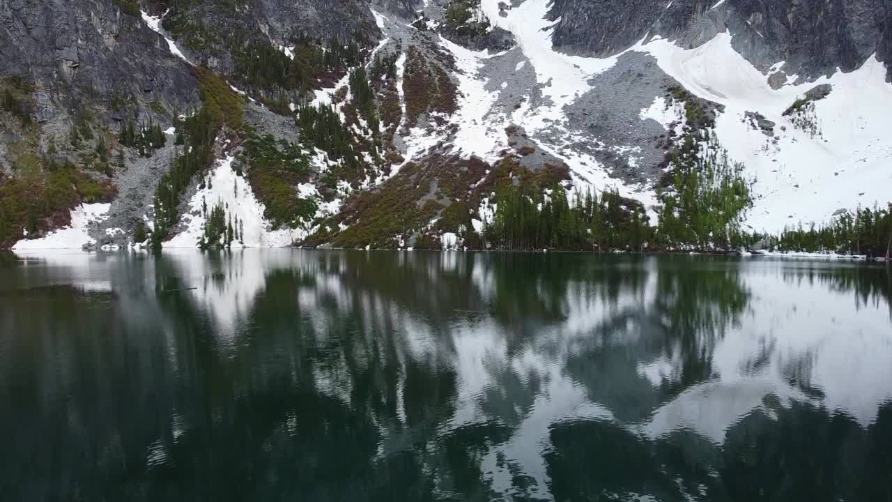 Low flying drone footage of Colchuck Lake, it's turquoise water, and the reflection of Aasgard Pass near The Enchantments in Washington's Cascade Mountain Range - Moss, trees, and snowy cliffside