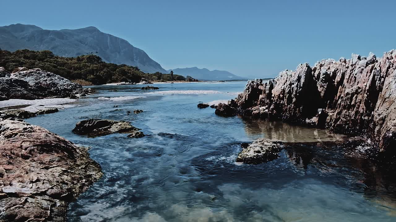 una hermosa escena con montañas en la lejanía y una laguna cristalina que fluye sobre rocas y arena en un entorno de estuario marino en hermanus, sudáfrica.