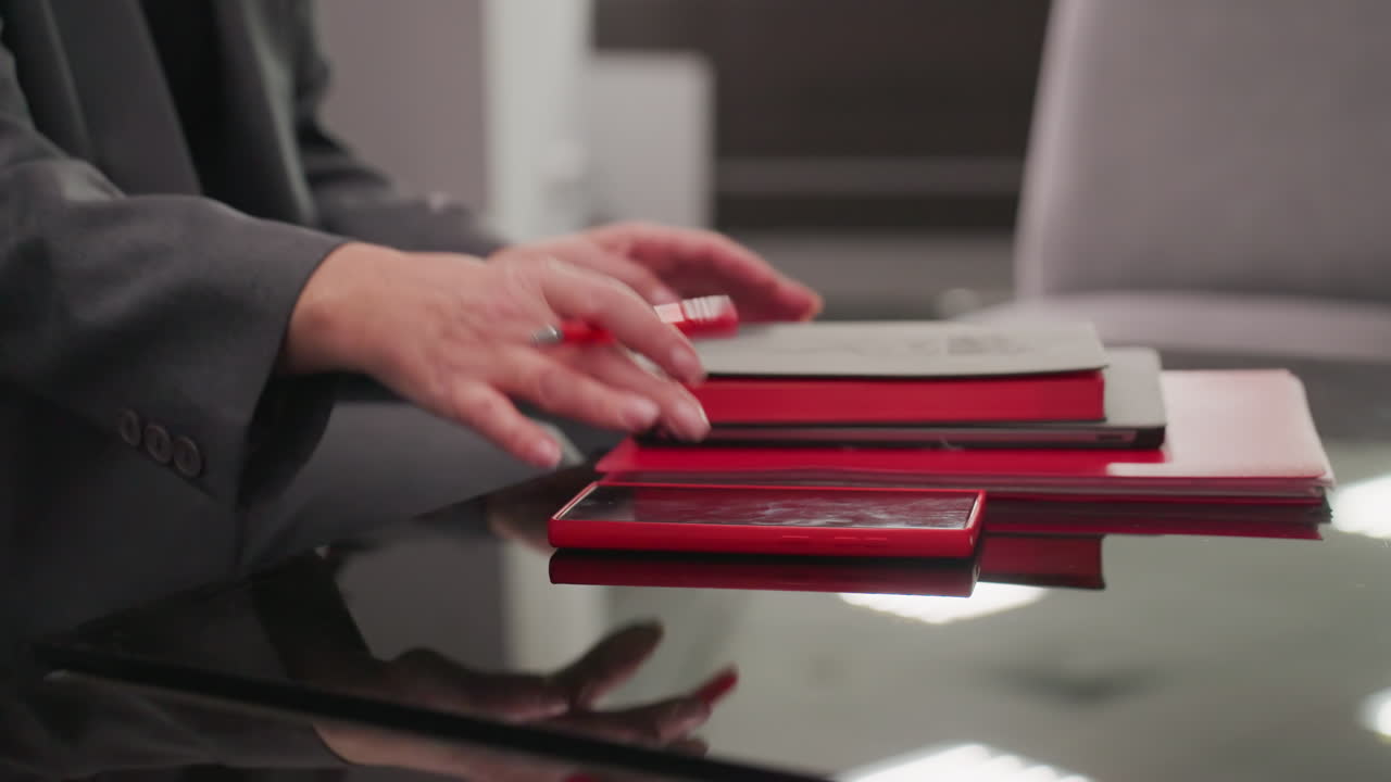 Close-up of businesswoman's hand resting on stack of red notebooks, smartphone, and papers on glass table. Female professional organized and focused, showcasing work environment and productivity