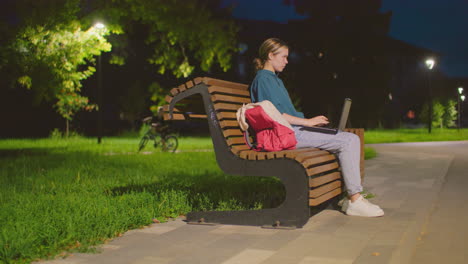 side view of woman sitting on outdoor bench at night, using laptop, illuminated by soft light, red backpack rests beside her on bench, with lush greenery and bicycle in background