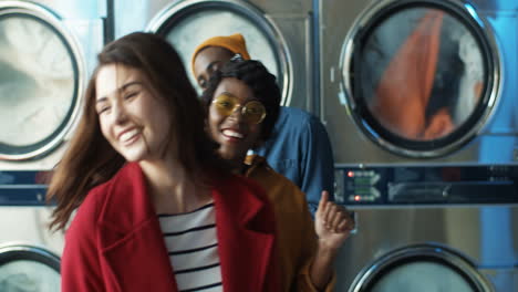 young blurred multiethnic joyful stylish girls and guy having fun and dancing in laundry service room while machines washing on background