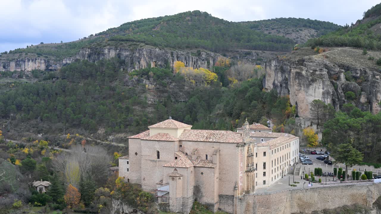 Perched on a cliffside in Cuenca, Spain, the historic Parador de Cuenca (a former convent) overlooks a gorge surrounded by hills with vibrant autumn foliage.