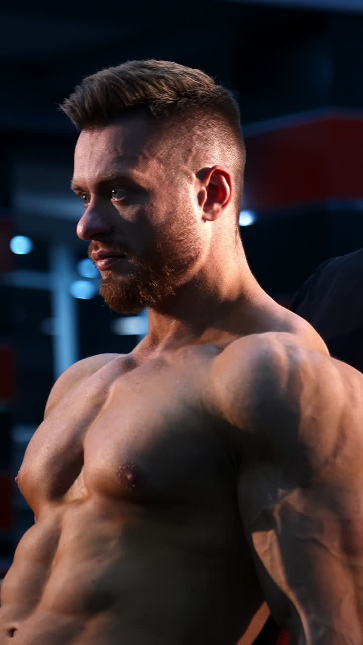 Shirtless bodybuilder in gym. Young guy with muscular body is lifting dumbbells. Sportsman practicing at sports center while sitting. Hard strength workout. Profile view. Vertical video