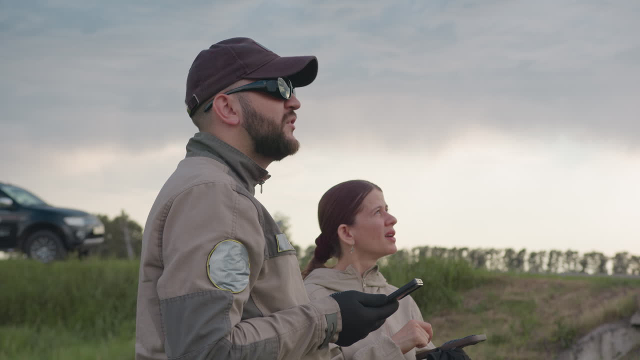 Bearded man in black cap and shades lifts smartphone skyward while woman checks tablet, both in khaki uniforms assessing flight trajectory under clouds near parked pickup along grassy embankment