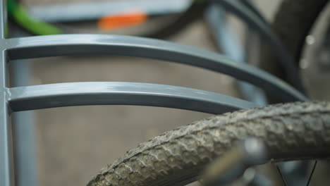 close-up of bicycle tire secured in metal bike rack with focus on textured wheel and metal frame, background shows another blurred bicycle parked nearby