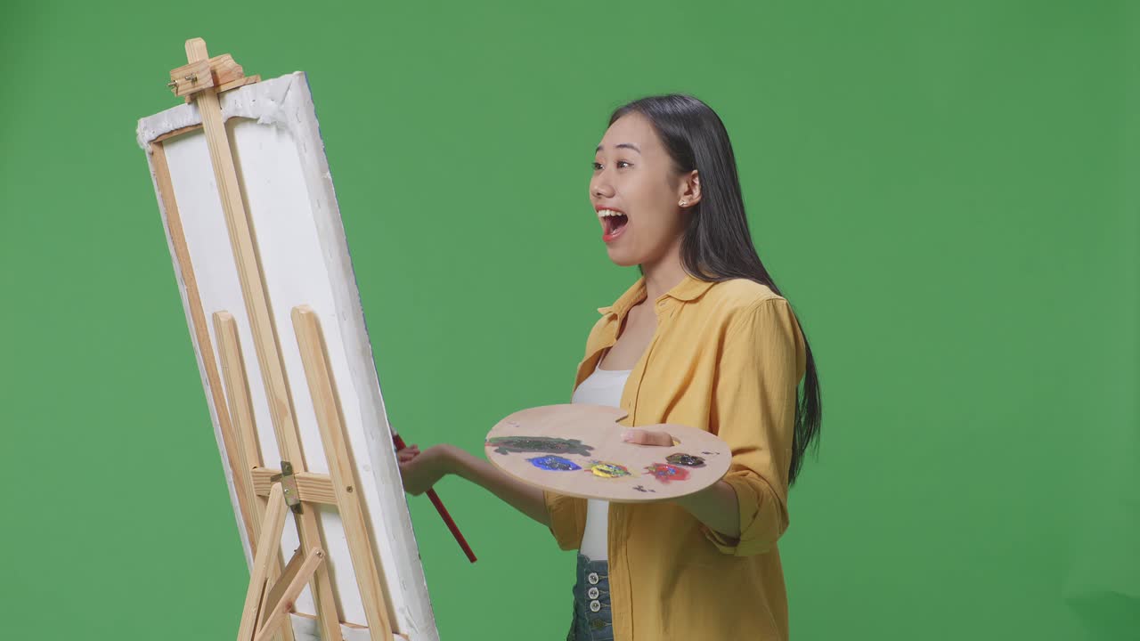 아시아 여성 예술가가 캔버스 위의 그림을 보고 와우라고 말하면서 녹색 스크린 스튜디오에서 오일 페인트와 브러쉬로 그림을 그리고 있습니다.