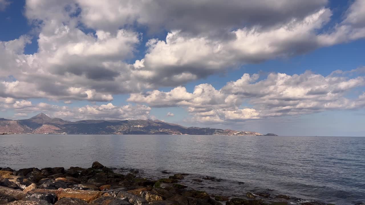 Close up of rugged rocky beach in northern crete greece with calm ocean and mountains under sunny blue sky with clouds