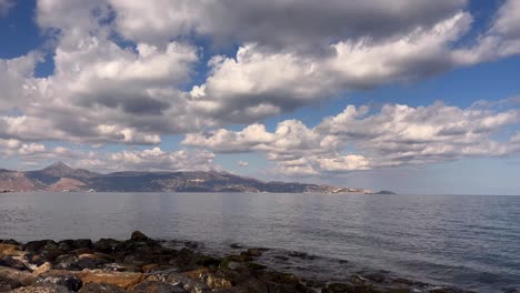 close up of rugged rocky beach in northern crete greece with calm ocean and mountains under sunny blue sky with clouds