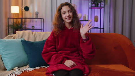 a young woman in a red hoodie sits on a couch and waves