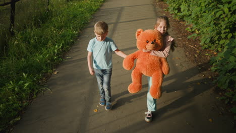 little girl walking on tarred path hugging large orange teddy bear while her younger brother gently holds teddy hand and looks back at something in background