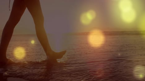 Glowing-yellow-spots-of-light-against-mid-section-of-a-woman-walking-on-the-beach