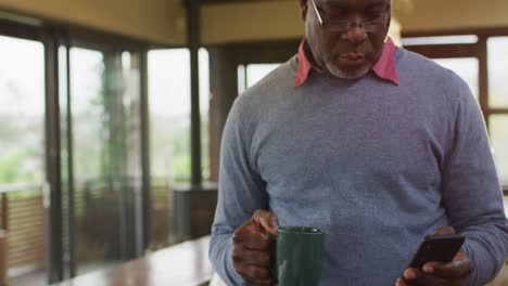 African-american-senior-man-at-counter-in-kitchen-using-smartphone-and-drinking-coffee