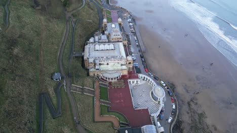 Seitenansicht-Aus-Der-Luft-Von-Der-Spitze-Des-Leerstehenden-Scarborough-Spa-Mit-Wunderschönem-Stadtbild-Im-Hintergrund-Und-Einem-Strand-Daneben-In-North-Yorkshire,-England