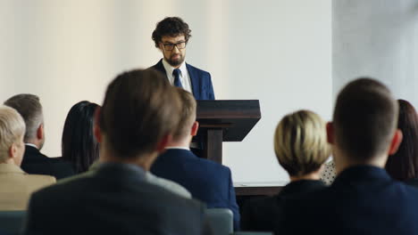 hombre de negocios caucásico con gafas y ropa formal hablando en una conferencia frente a gente de negocios