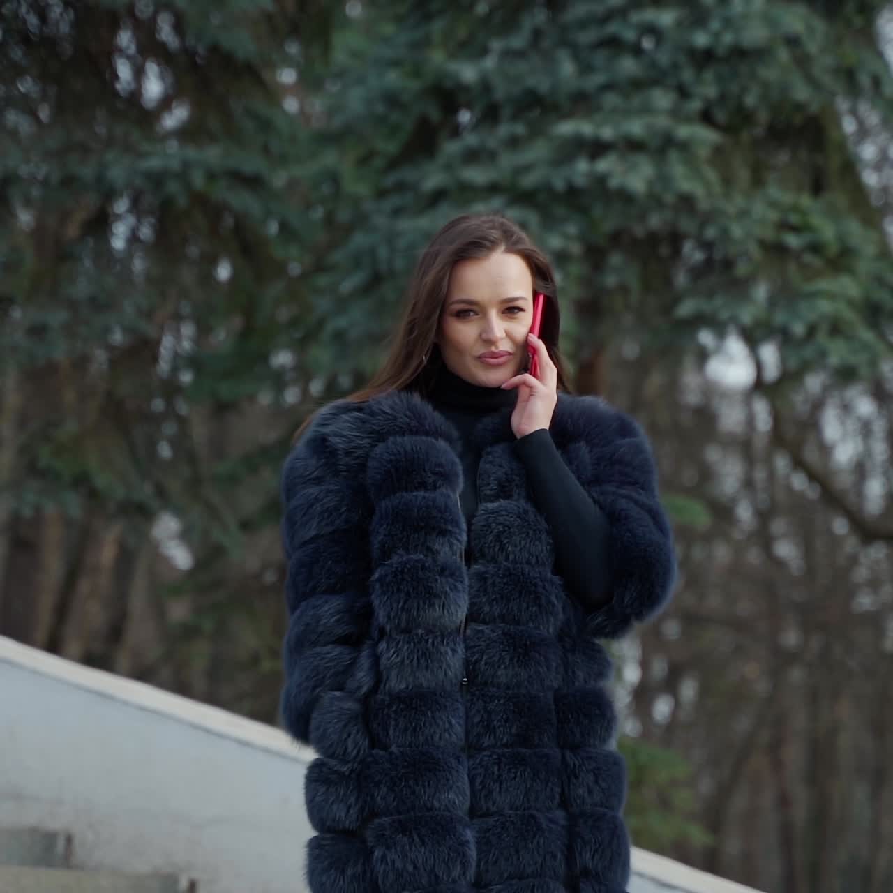 Luxury girl with a phone in the park. Portrait of attractive lady in fur coat walking in the park and talking the mobile phone. Slow motion.