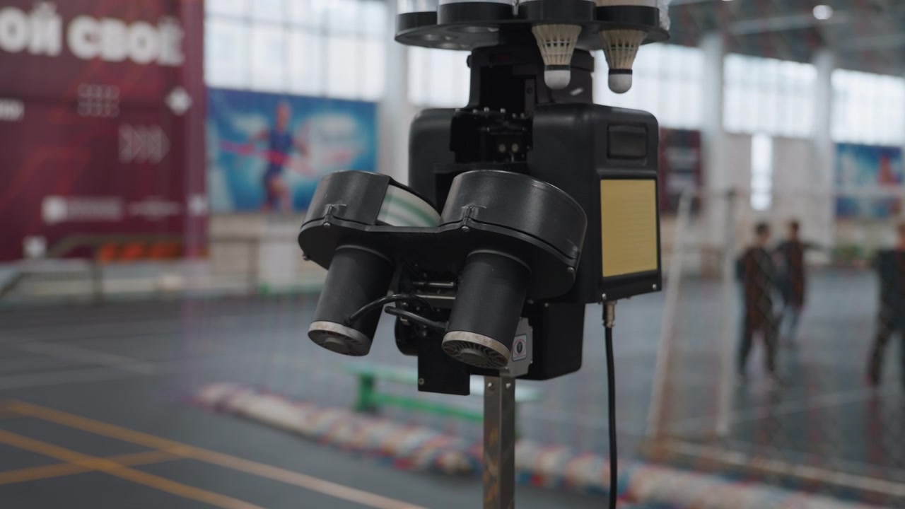 Máquina lanzadora de bádminton en pista interior, primer plano del alimentador automático de volantes con jugadores borrosos practicando al fondo, pabellón deportivo de techo alto, pancartas y suelo sintético, ejercicios de velocidad.