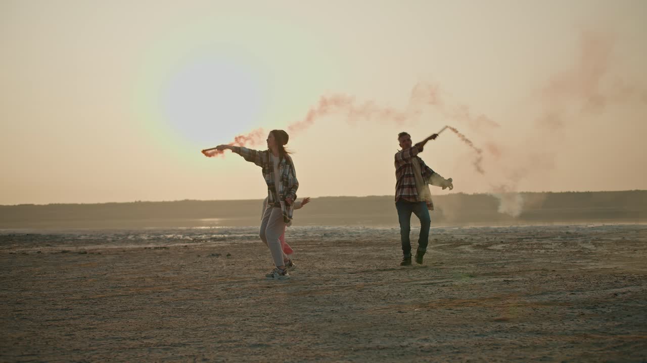 una feliz chica morena en una camisa a cuadros verde junto con su marido y su hija pequeña juegan con fuegos artificiales que emiten humo verde y rojo en la orilla de un mar desierto durante sus vacaciones fuera de la ciudad en la noche de verano