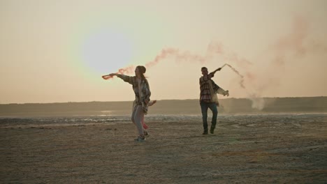 a happy brunette girl in a green checkered shirt together with her husband and little daughter play with fireworks that emit green and red smoke on the shore of a deserted sea during their vacation outside the city in the summer evening